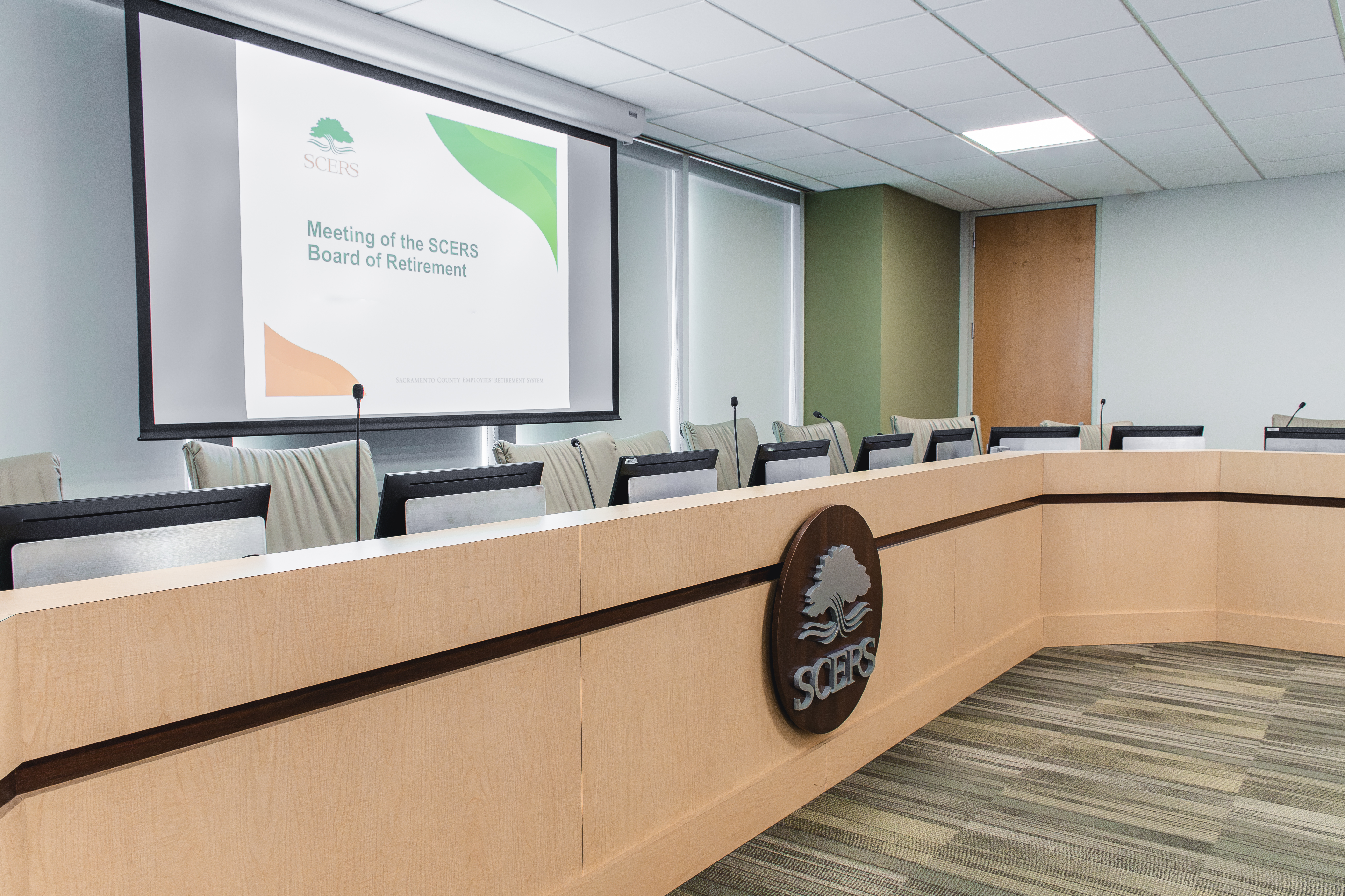 Empty boardroom with long wooden table, equipped with microphones and chairs. A screen displays 